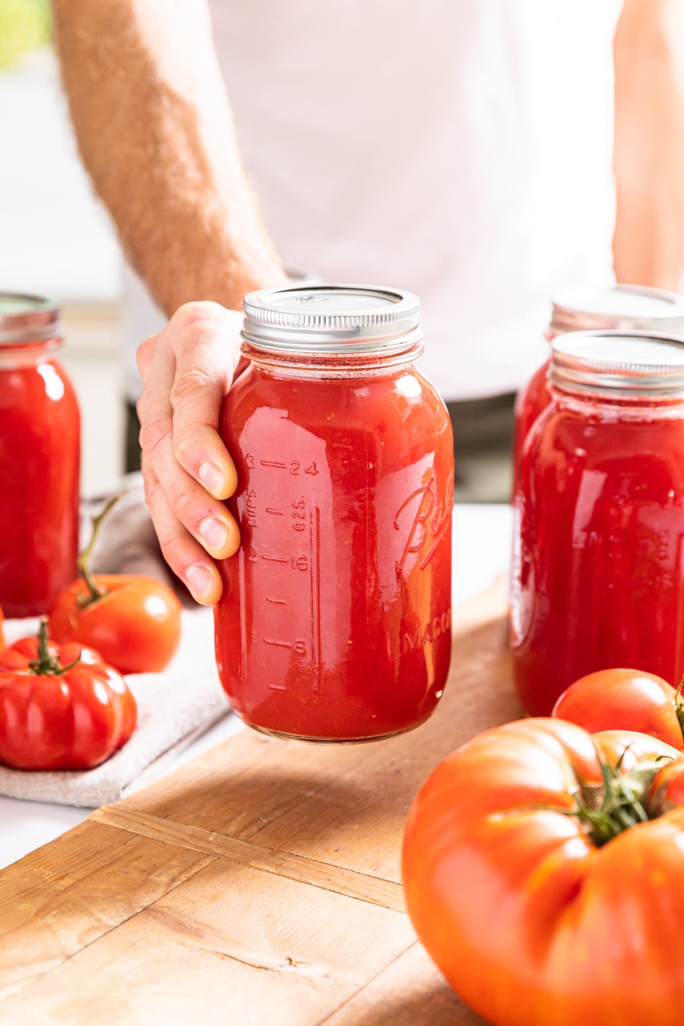 Hand holding glass jar filled with red colored tomato juice after being canned with wood board underneath
