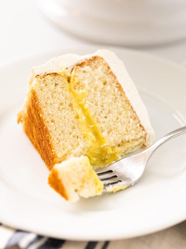 Slice of white layer cake filled with yellow pudding interior with white frosting with fork scooping off piece all on white plate