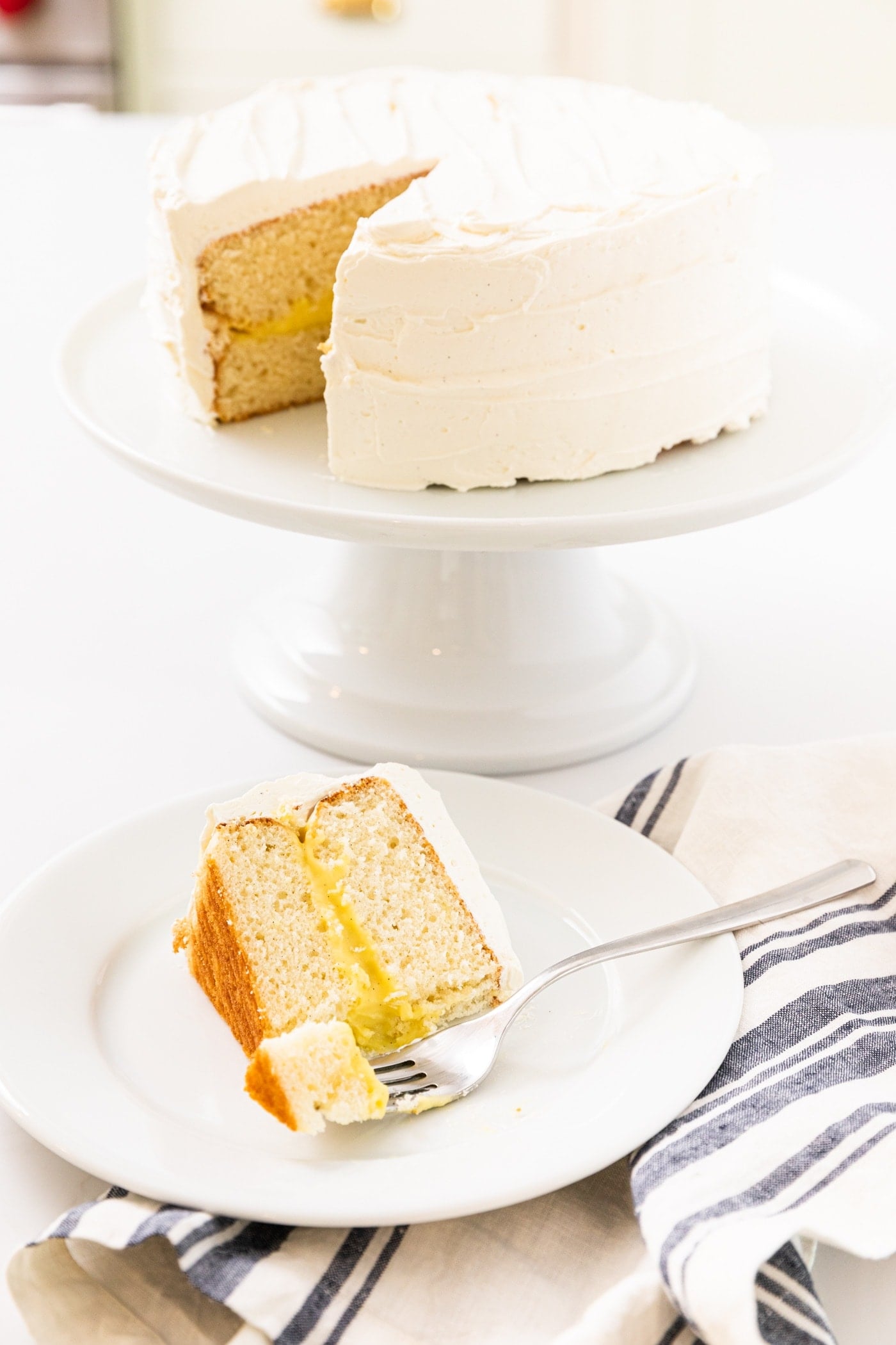 White frosted layer cake sitting on white cake stand in background with slice of cake sitting on white plate with fork in front all one white countertop
