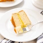 Slice of white cake with fork scooping off piece topped with white frosting and yellow colored middle layer sitting on white plate on white countertop
