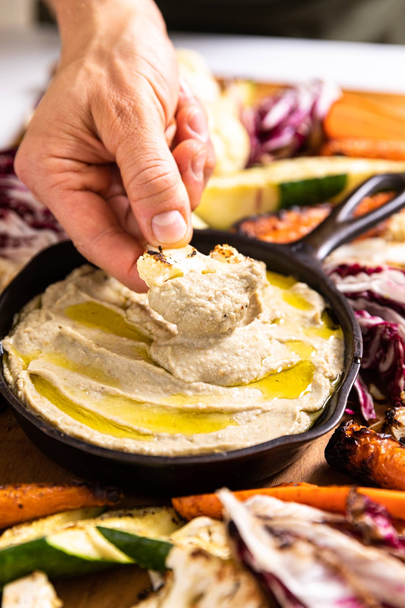 Hand holding piece of grilled cauliflower dipping into light brown colored Baba ganoush drizzled on top with olive oil and sitting in black cast iron skillet