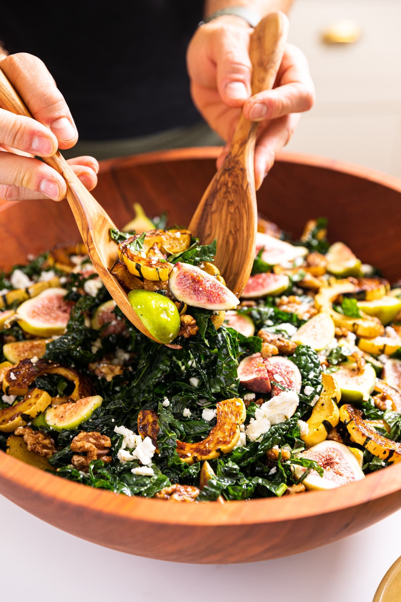 Two hands holding wooden tongs dipping salad out of a wooden bowl with pieces of kale and fig and squash