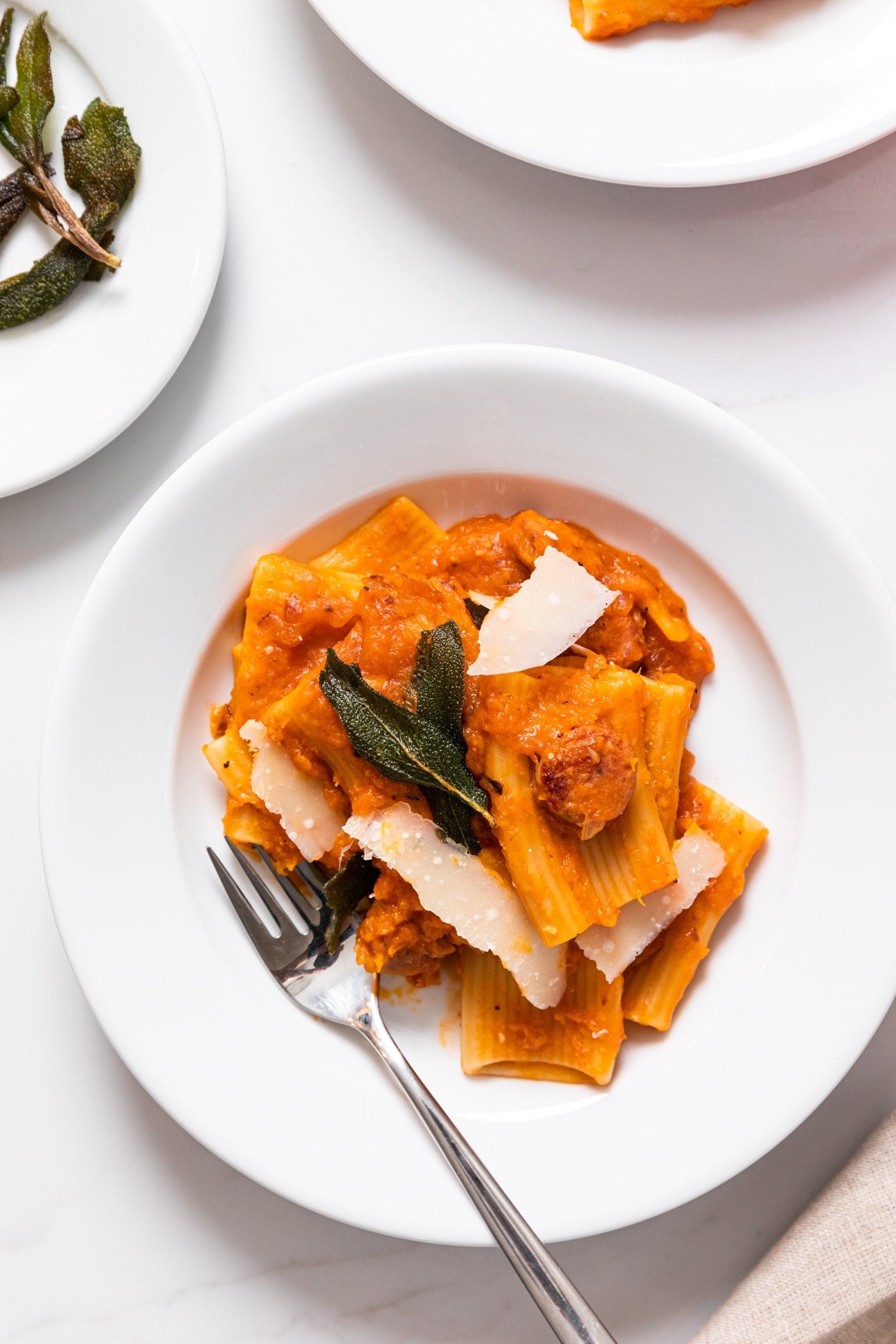 Top down view of white plate filled with large pieces of tubular pasta topped with fried sage on white countertop