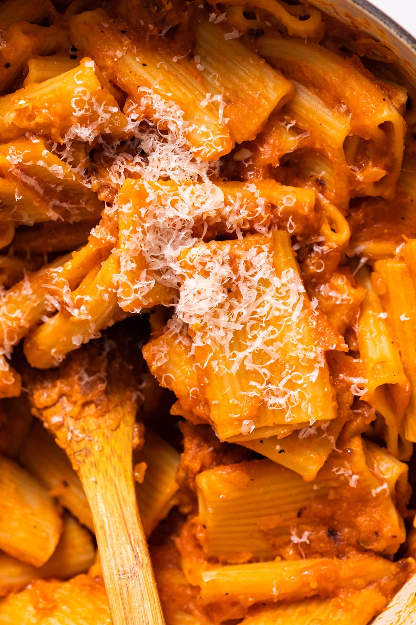 Top down view of orange colored tubular shaped pasta covered with sauce and topped with cheese with wooden spoon in the middle