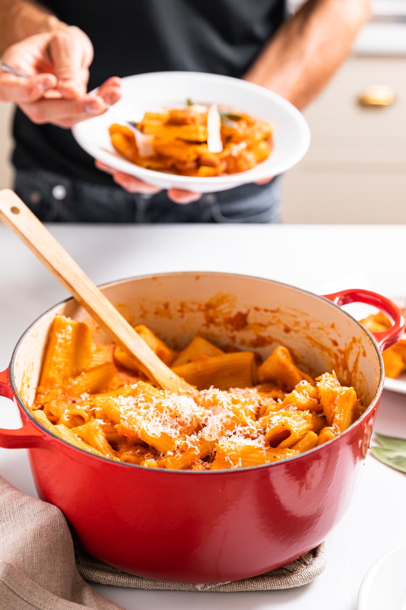 Large red pot sitting on white countertop with wooden spoon inside with hand holding plate filled with pasta