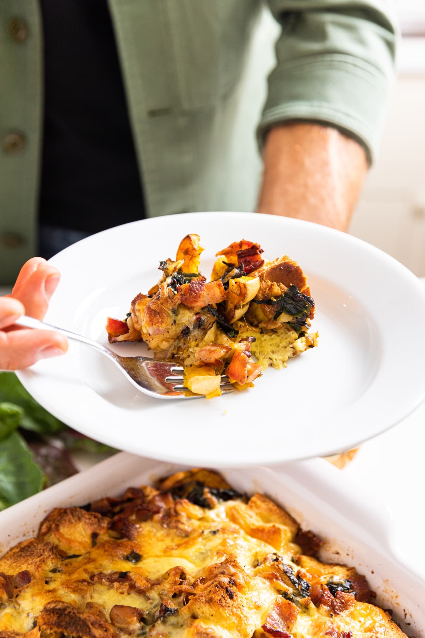 Hand holding white plate filled with scoop or squash strata with fork taking out a piece with rest of strata in white casserole dish below
