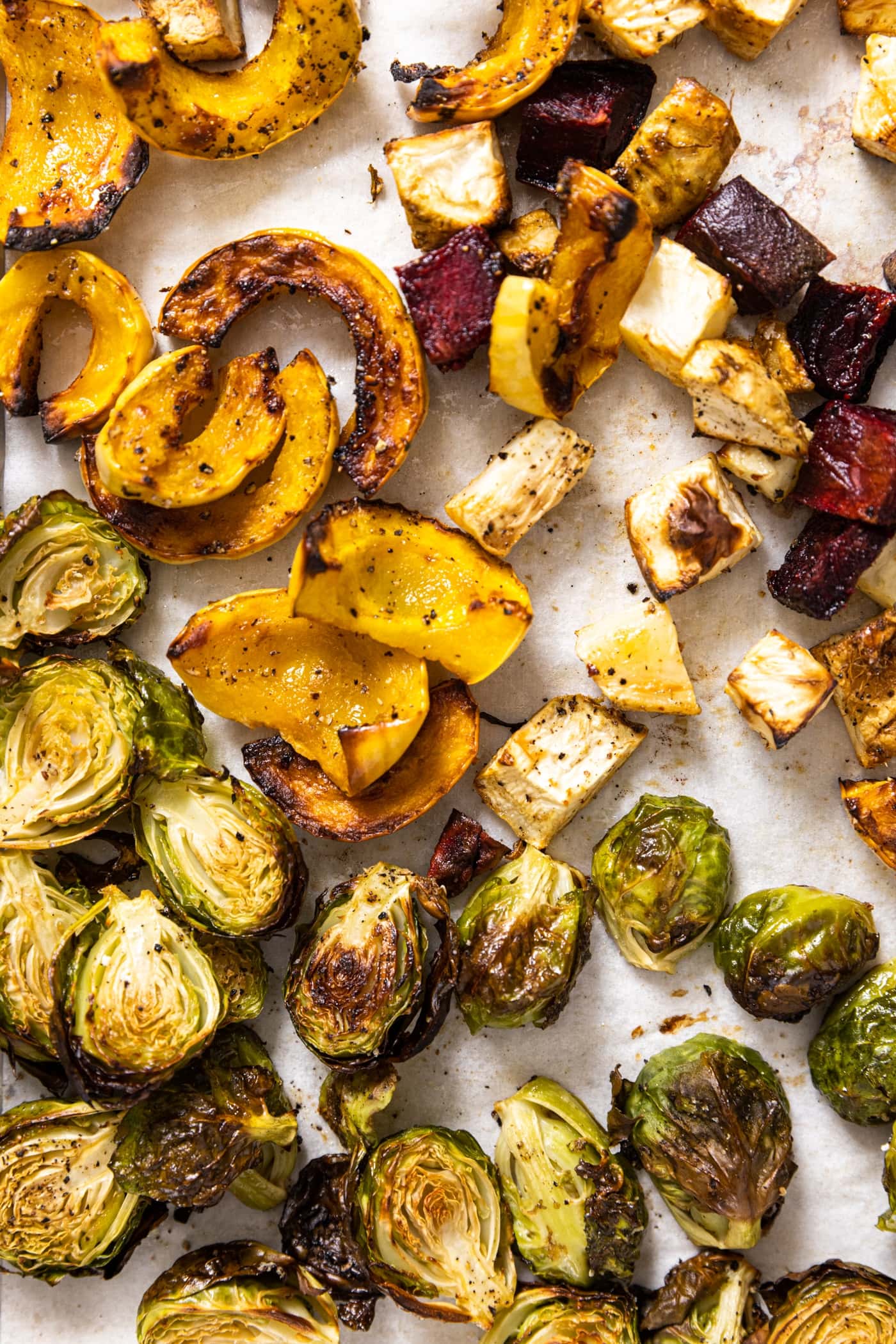 Top down view of baking sheet with white piece of parchment with roasted vegetables all over the top including squash, beets, and Brussels sprouts