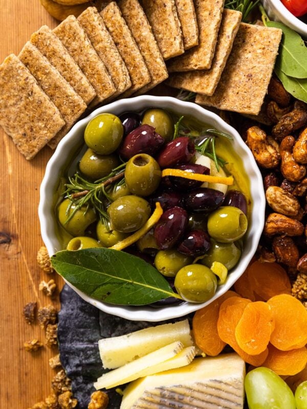 Top down view of small white bowl filled with black and green marinated olives sitting on wood board with cheese and crackers and dried fruit