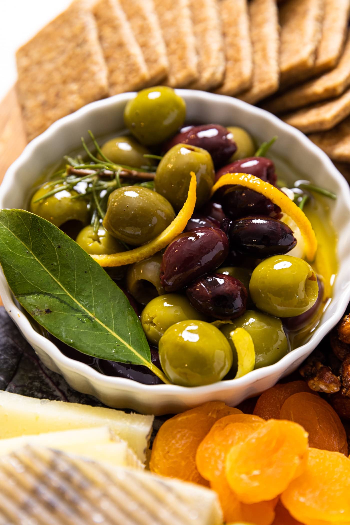 Small white bowl filled with marinated olives with slices of citrus zest sitting on wood board with crackers and dried apricots