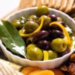 Small white bowl filled with green and black olives with slices of lemon zest with a bay leaf sitting on wood board