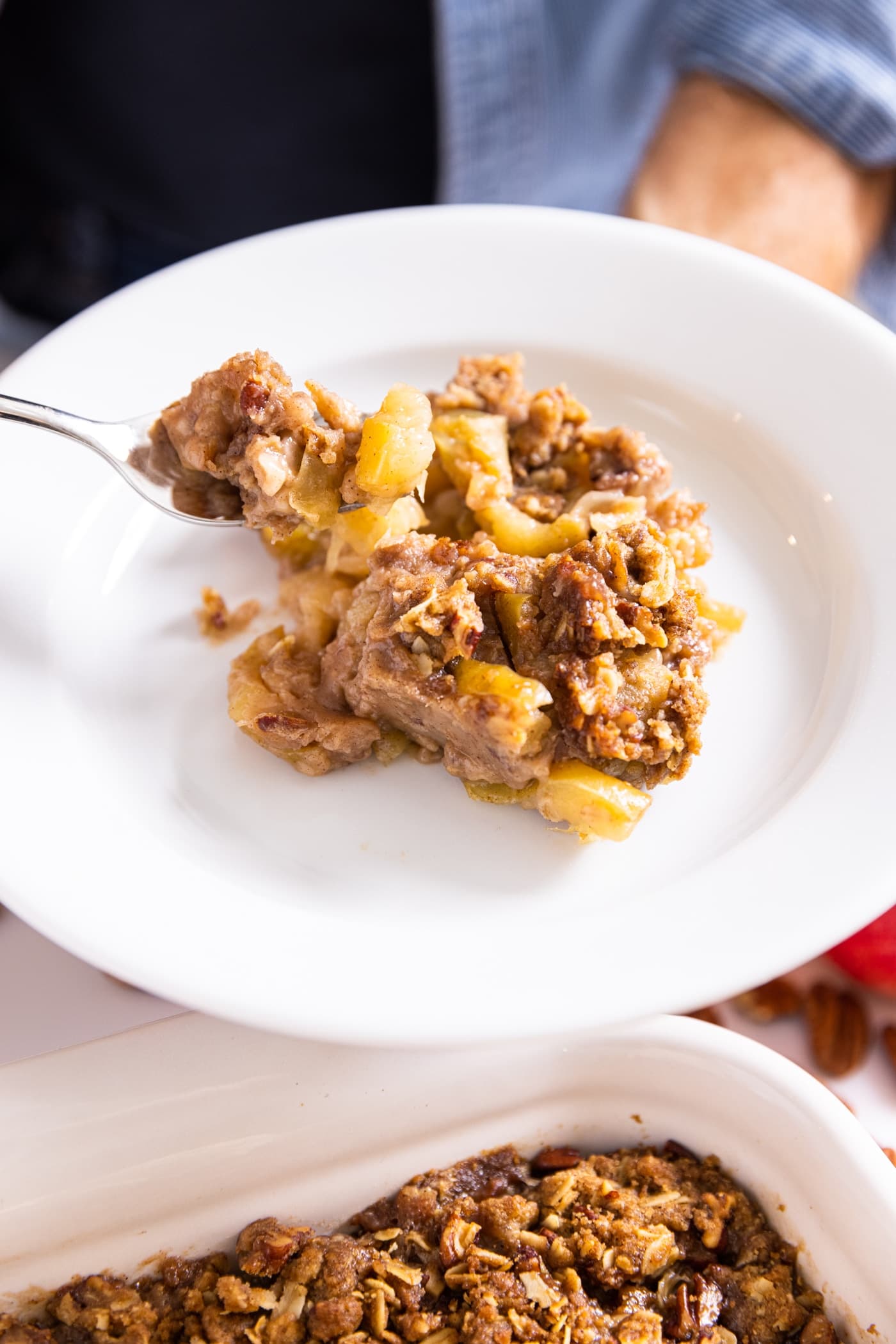 Fork holding bite of apple crisp with streusel topping on white plate with rest of crisp below
