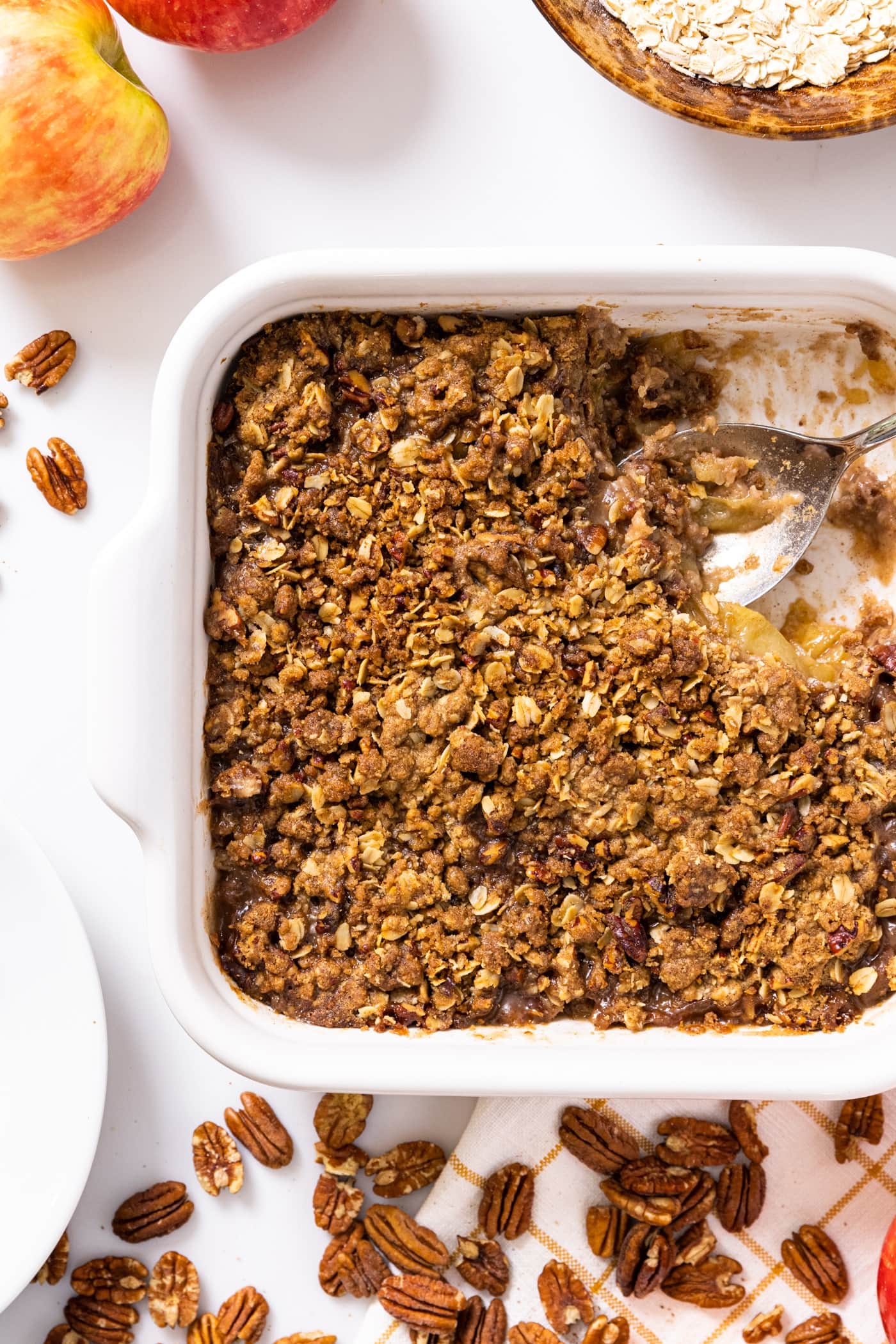 Top down view of white baking dish filled with brown colored apple crisp sitting on white countertop with pecans and apples all around