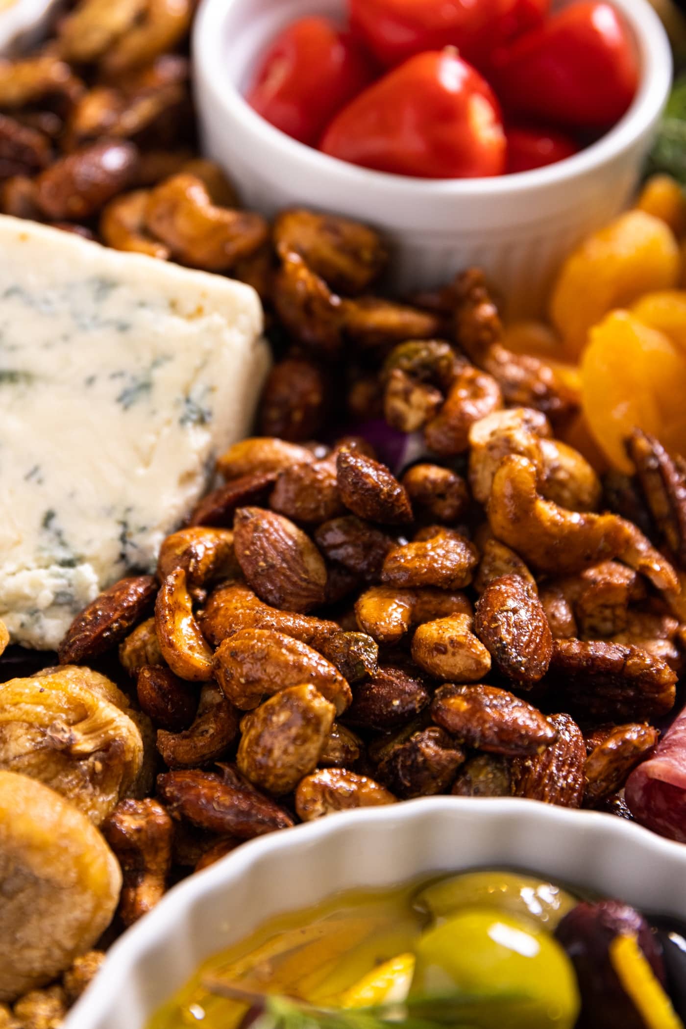 Close up view of spiced nut mix sitting on charcuterie board amongst red stuffed peppers, olives, dried fruit, and blue cheese
