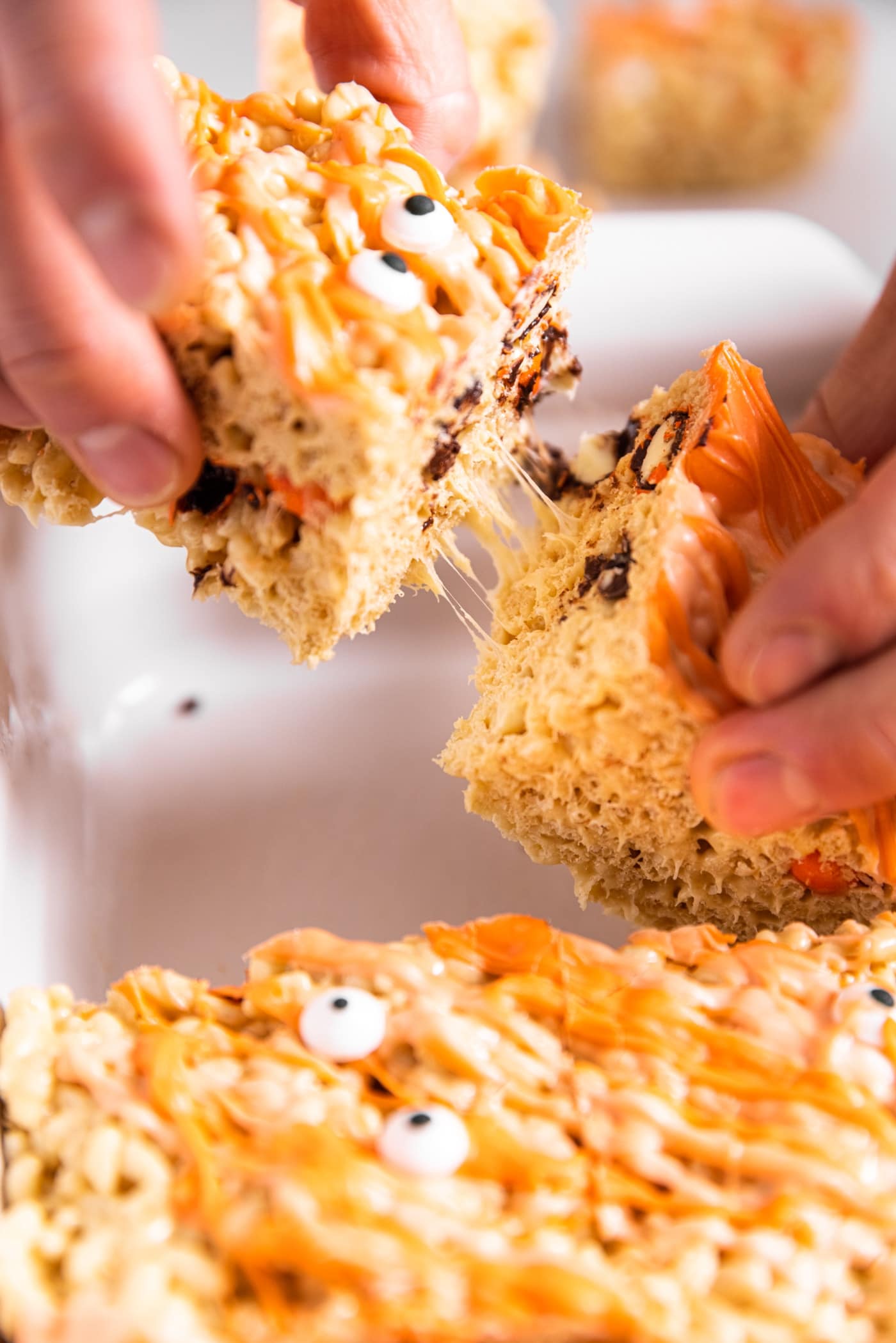 Two Rice Krispie treats being pulled apart by two hands with strings sticking together between the two with the rest of the pan
