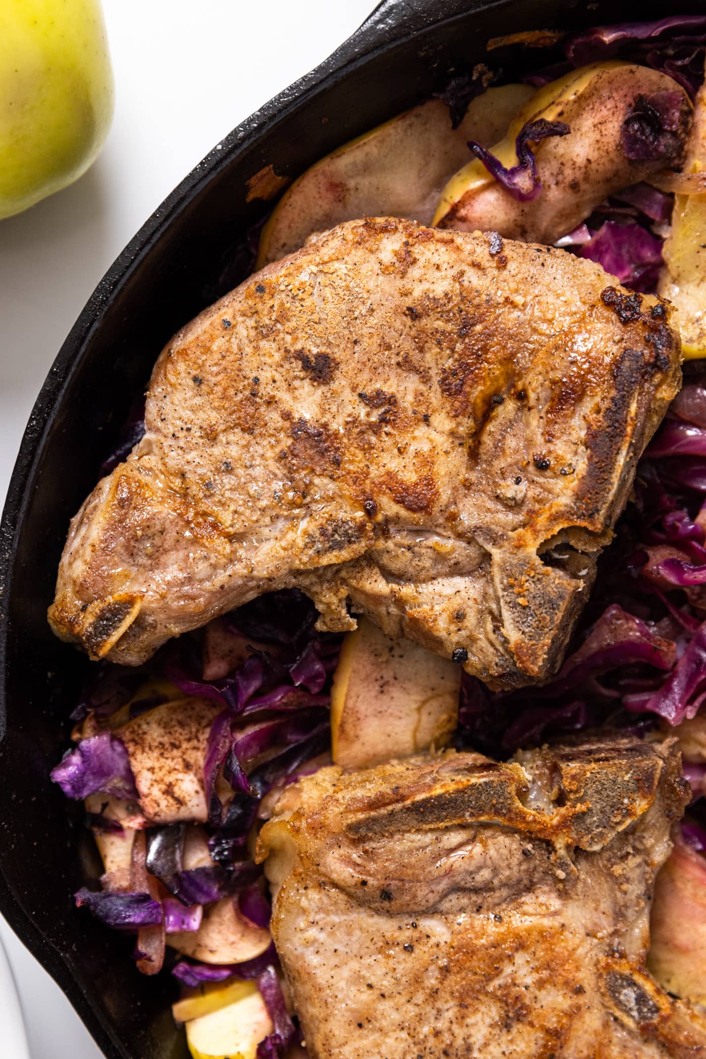 Top down view of cast iron skillet with red cabbage and apples underneath nestled pork chops with a crisp crust