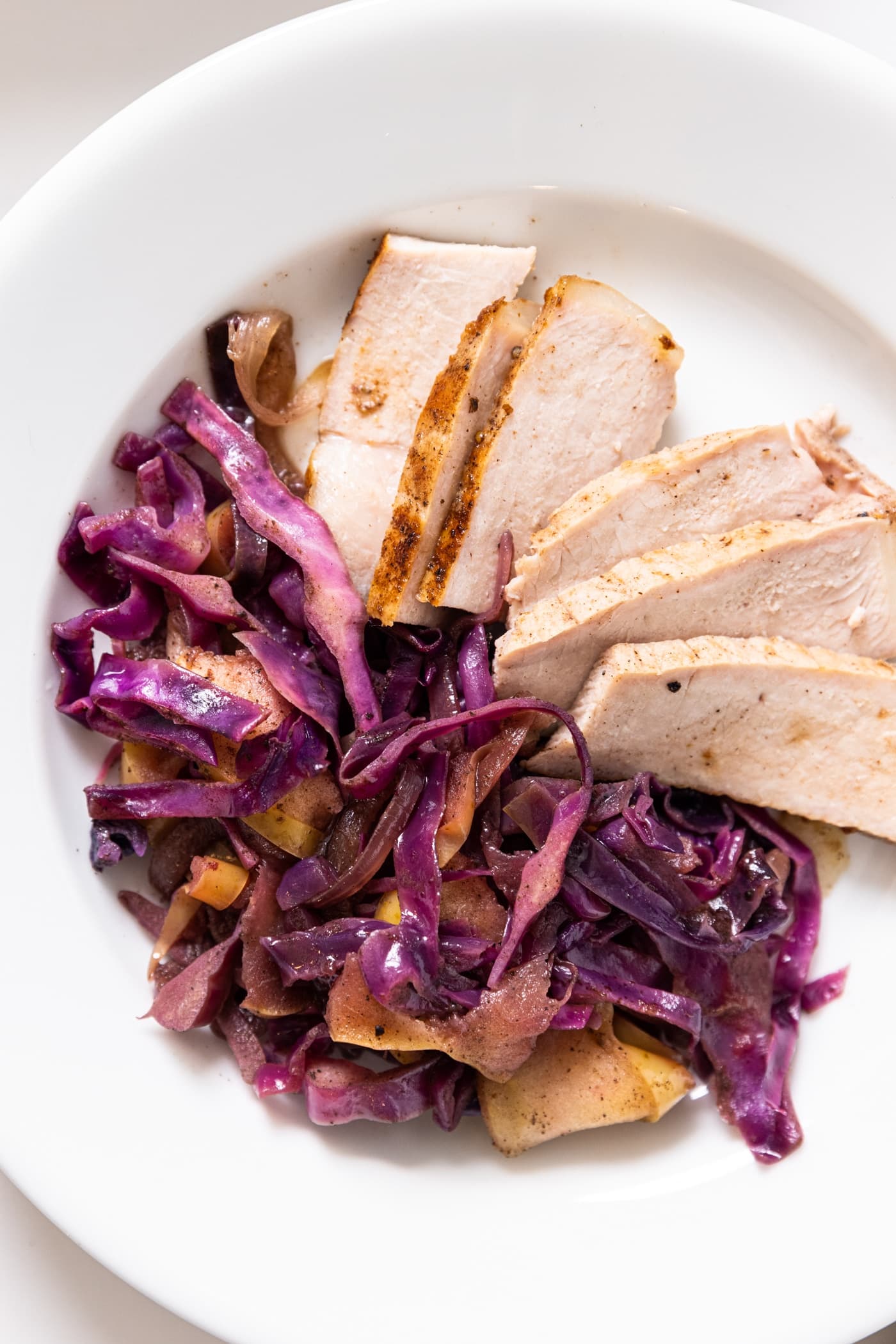 Top down view of white plate filled with slices of pork with red cabbage and apples on the side