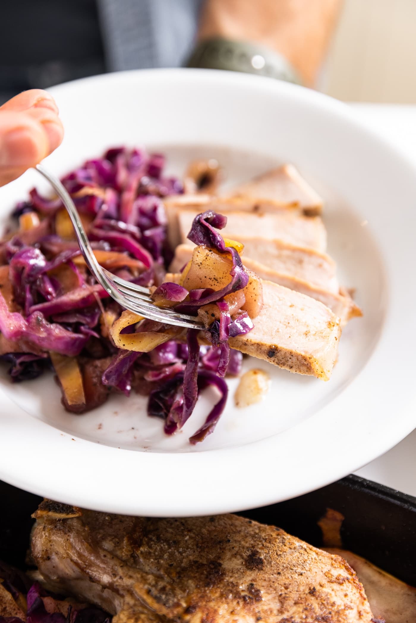 White plate filled slices of pork and a combination of red cabbage and apples with fork holding bite full