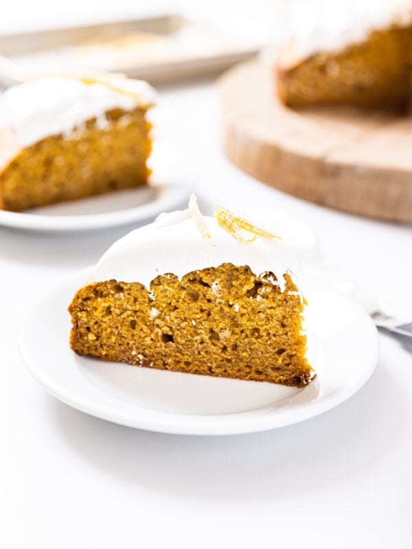 Slice of pumpkin cake sitting on white plate topped with white whipped cream with other slice and rest of cake in background