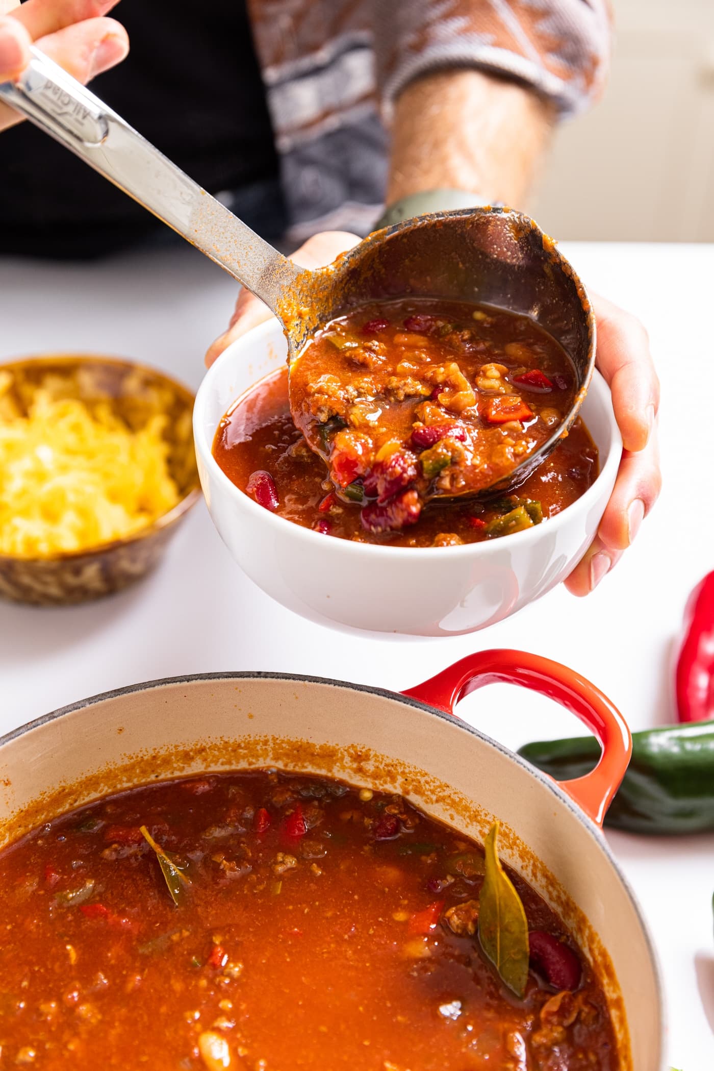 Hand holding white bowl pouring red colored chunky chili into bowl with rest of soup on white countertop