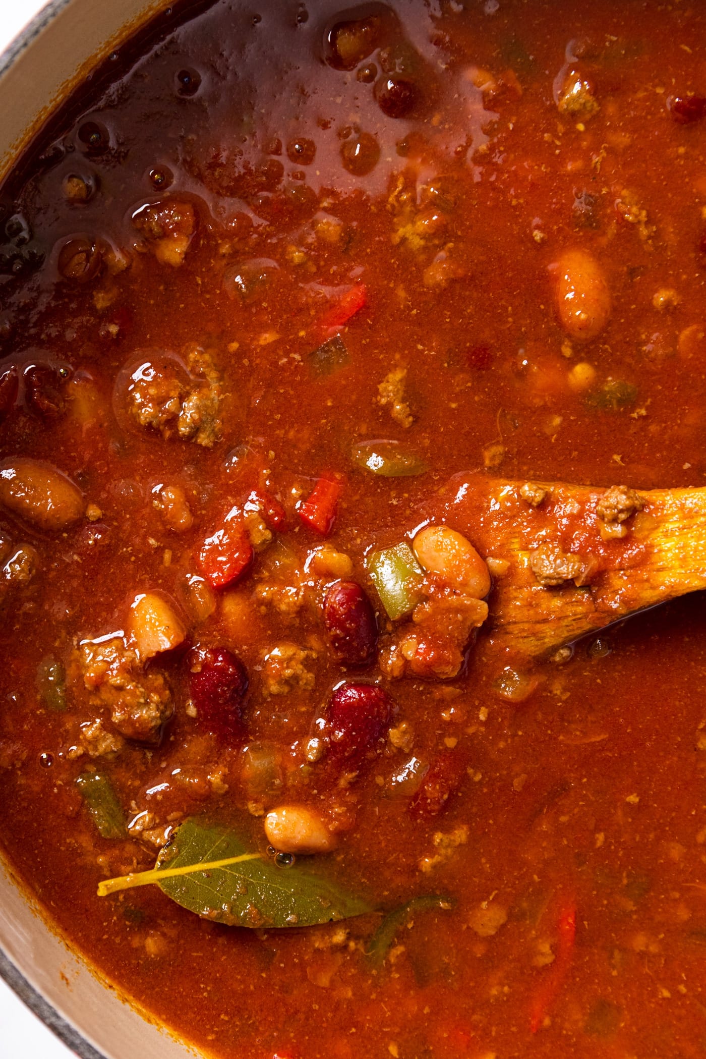 Top down view of big red kettle with wooden spoon in the middle of bright red chili with beans and meat and a bay leaf