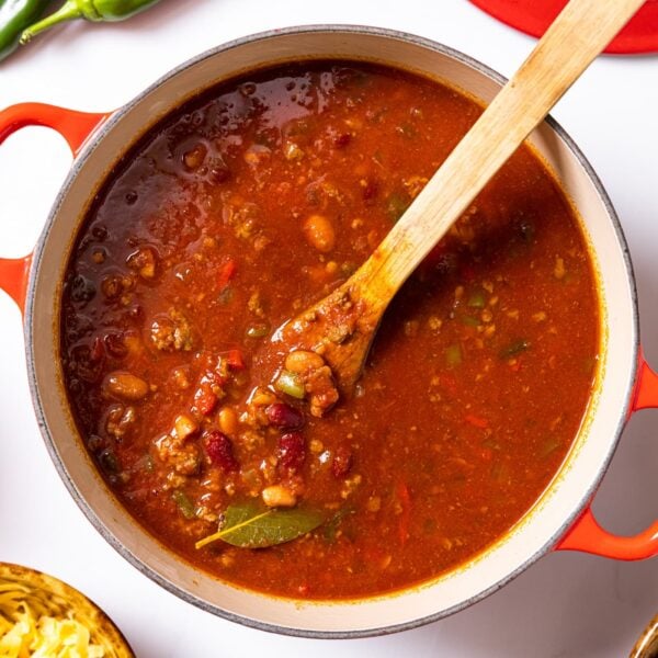 Top down view of red Dutch oven sitting on white countertop filled with red chili with peppers, crackers, and cheese in other bowls all around