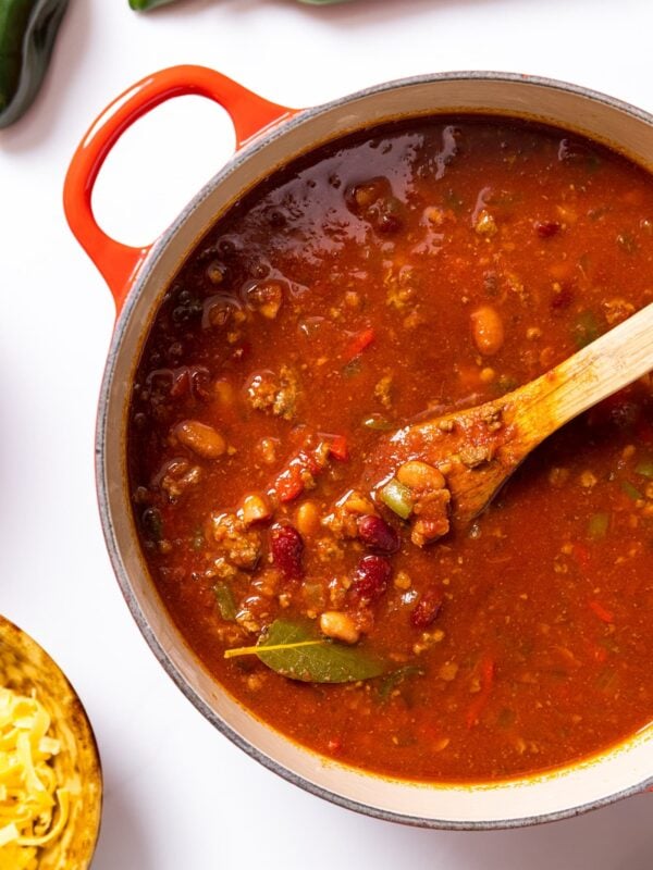 Top down view of large red kettle filled with red colored chili sitting on white countertop with green peppers and cheese around
