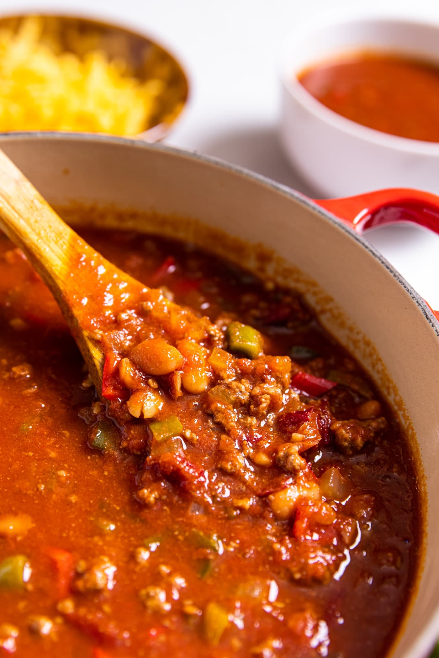 Large red Dutch oven sitting on white countertop with wooden spoon pulling up some of bright red chili showing the beans and meat and red sauce
