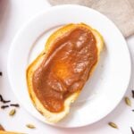 Top down view of white plate with slice of bread sitting on top with slathered brown pear butter one top with extra pears and spices on white countertop