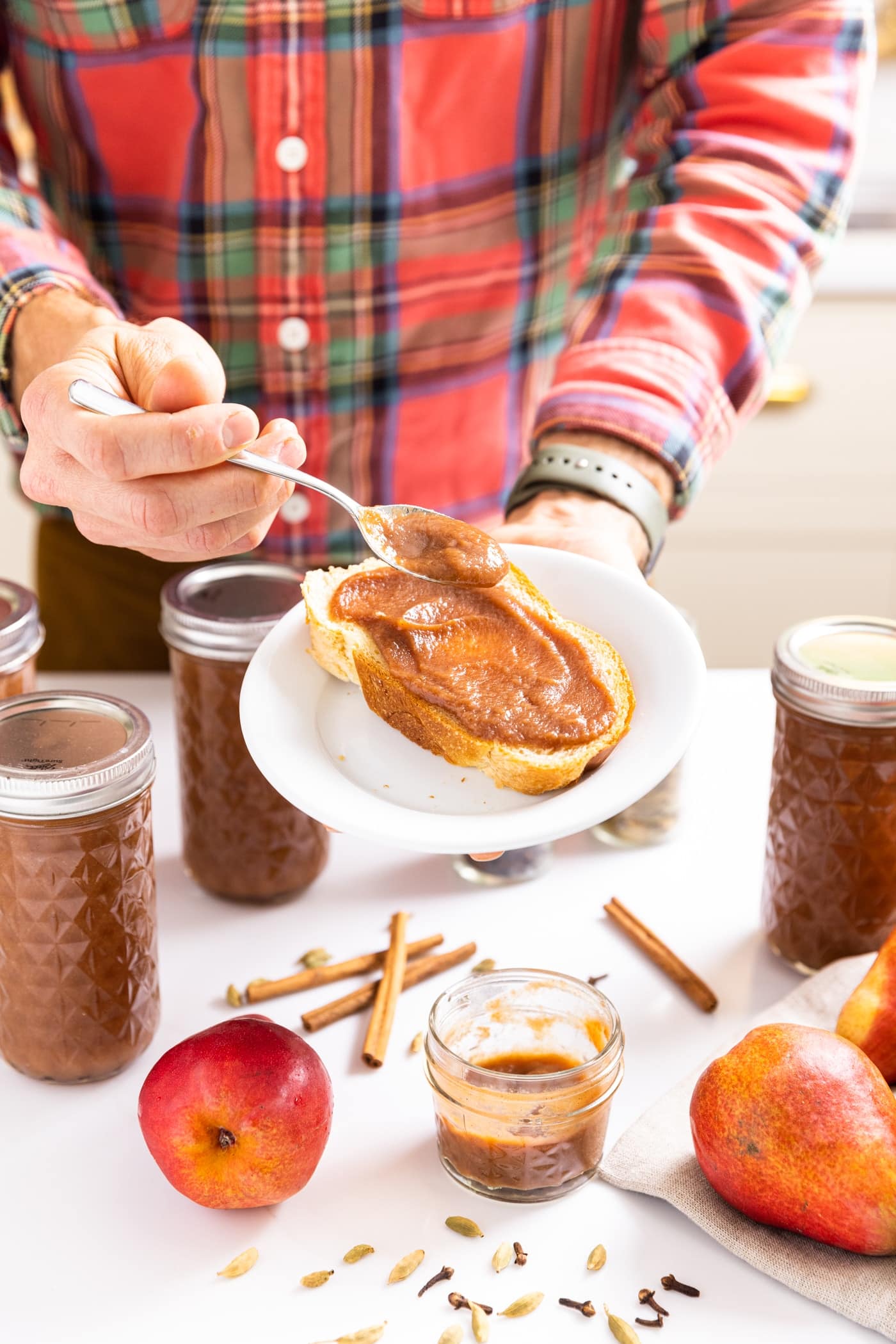 Two hands holding white plate with slice of bread on it with spoon spreading pear butter onto bread
