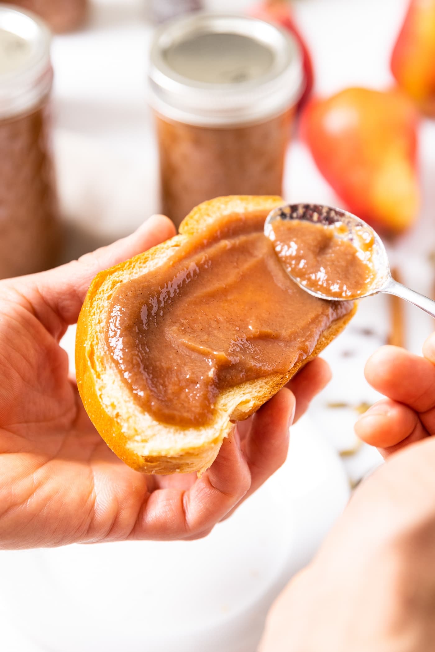 Hand holding slice of bread as pear butter is spread all across with glass jars filled with pear butter in background