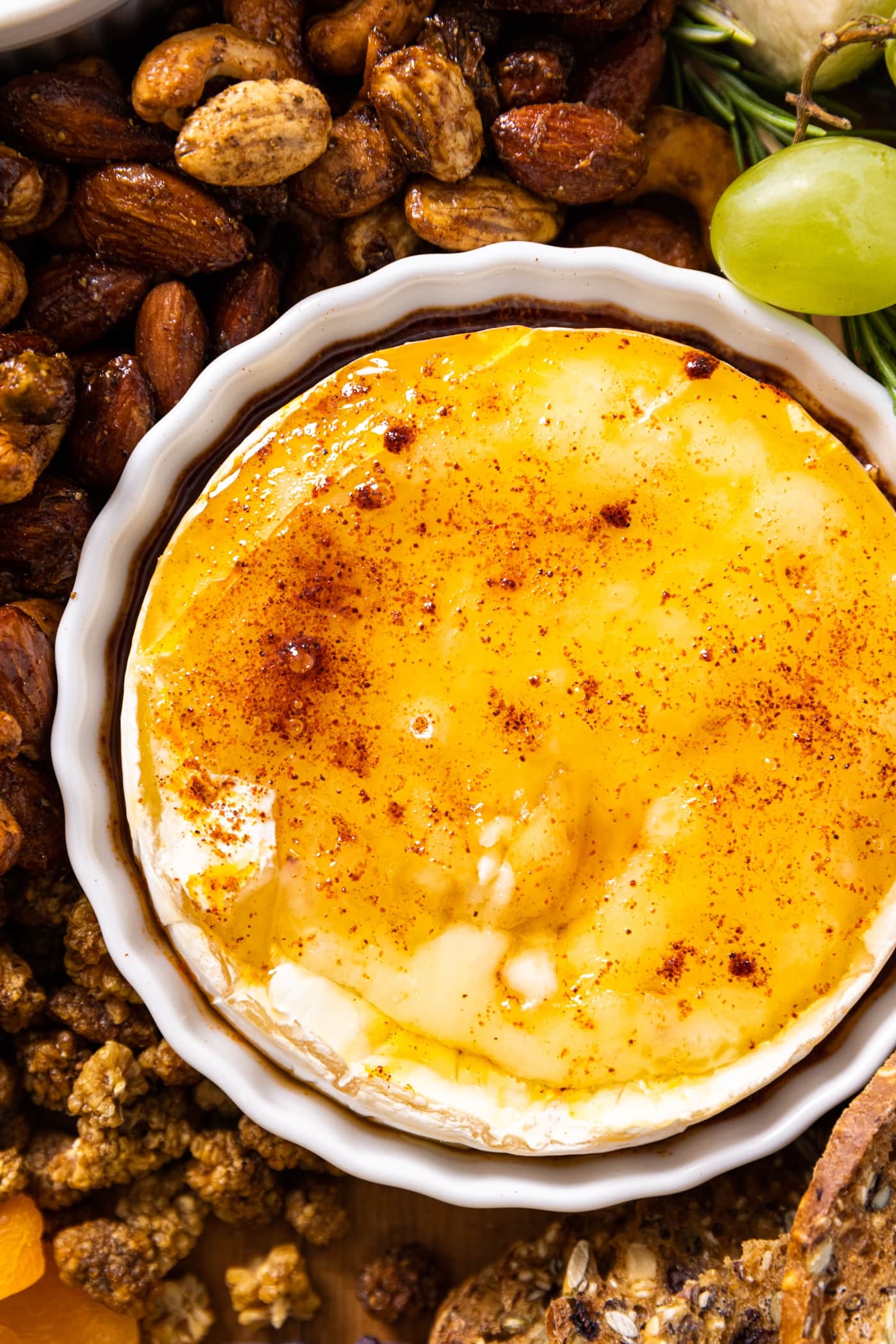 Top down close up view of baked Brie topped with honey and chipotle powder sitting in white bowl amongst spiced nuts and grapes