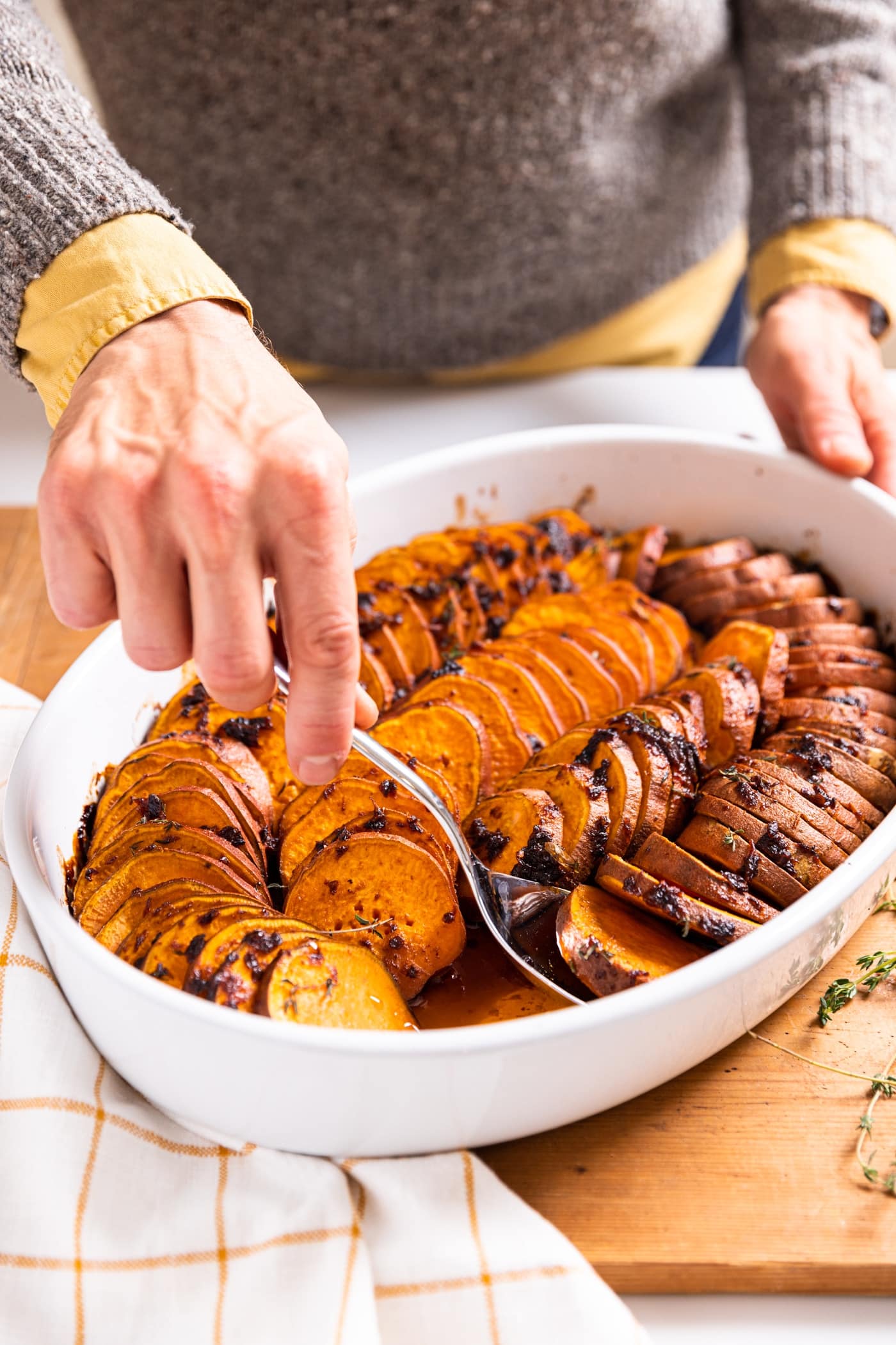 Hand scooping slices of sweet potatoes out of white serving dish sitting on wood board