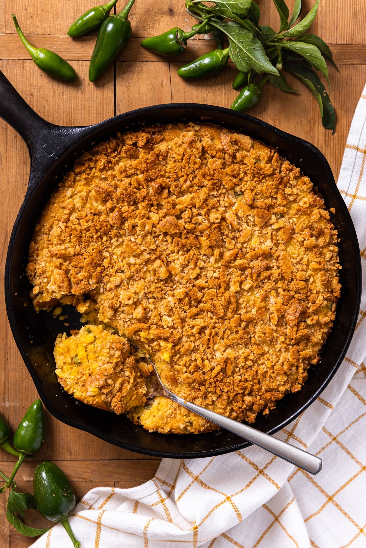 Large black cast iron skillet sitting on wood board filled with golden colored cracker crusted corn spoon bread with jalapenos all around