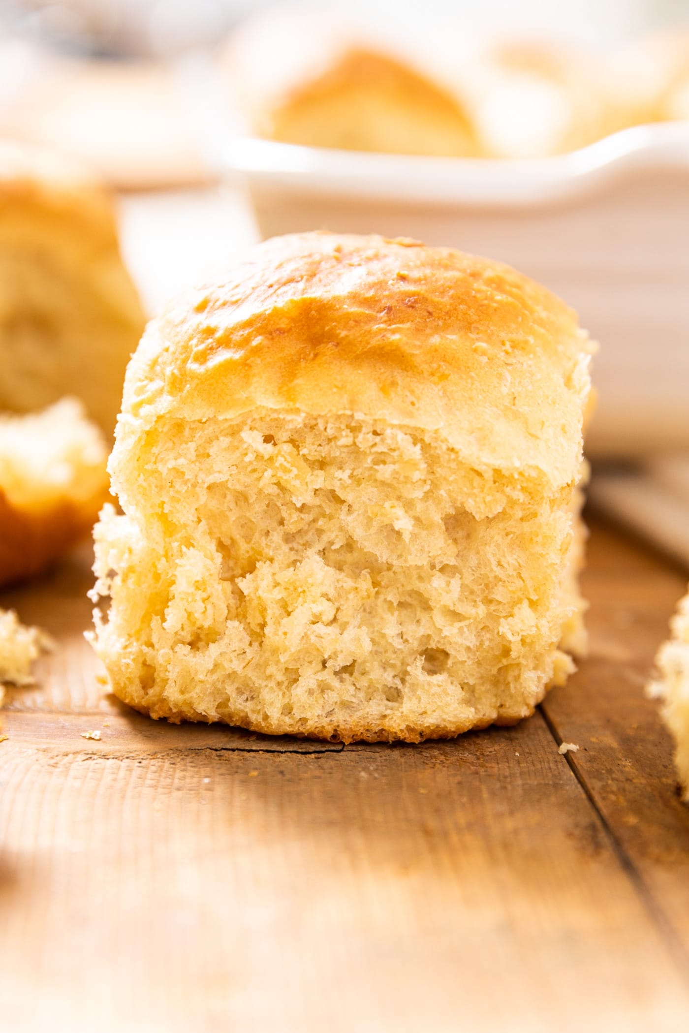 A single golden brown dinner roll sitting on wooden board with white baking dish in background filled with more dinner rolls