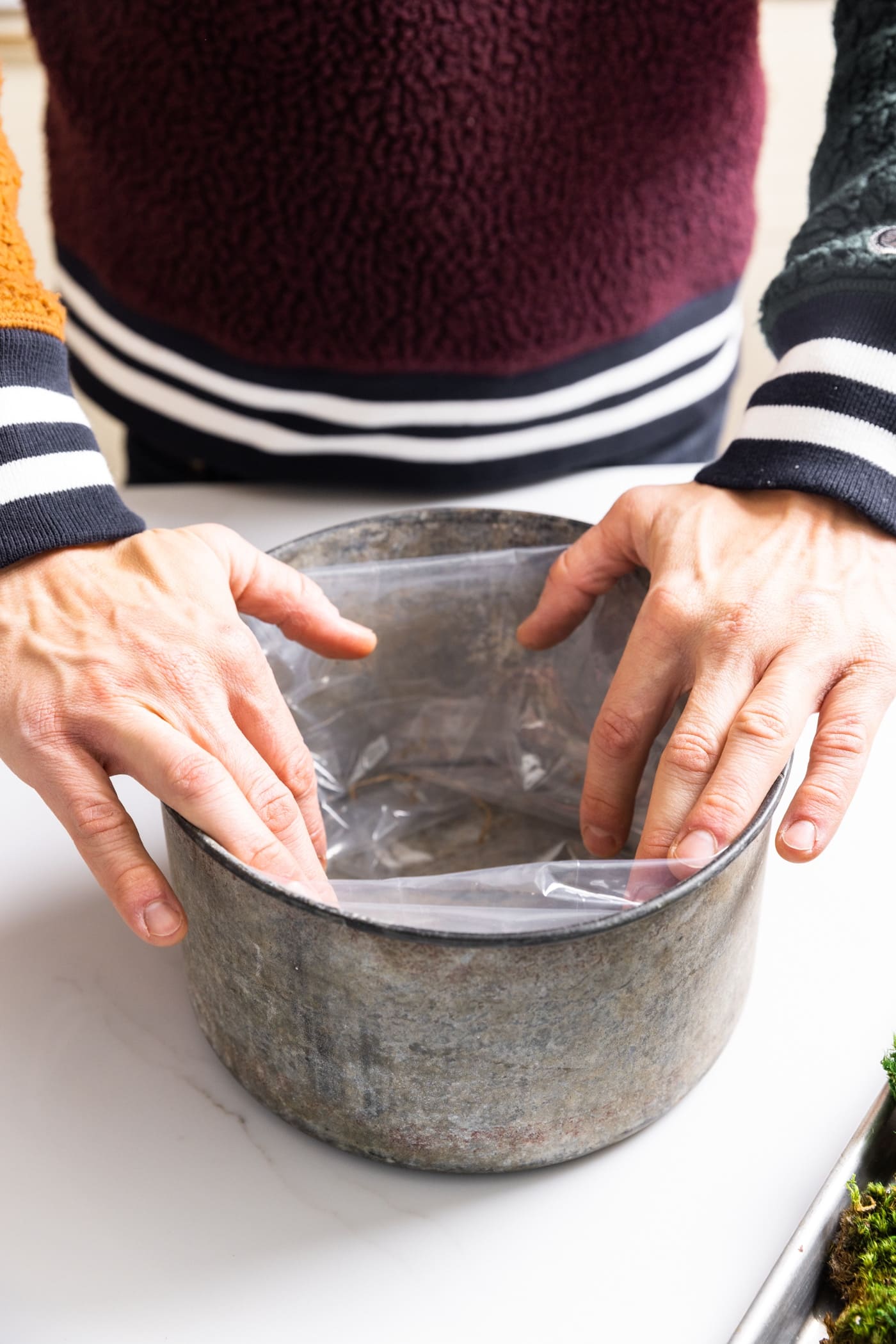 Two hands holding plastic around edge of galvanized metal container sitting on white countertop