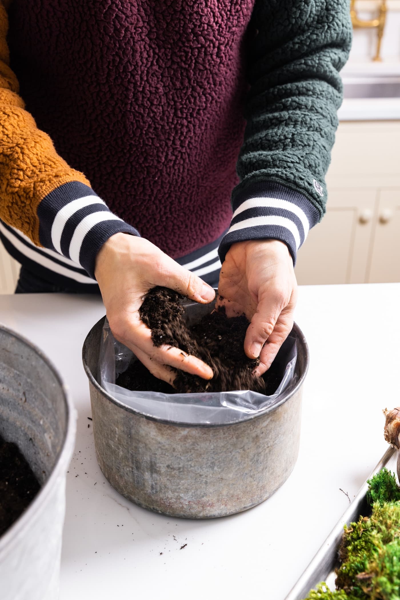 Two hands putting soil in a small galvanized container on white countertop