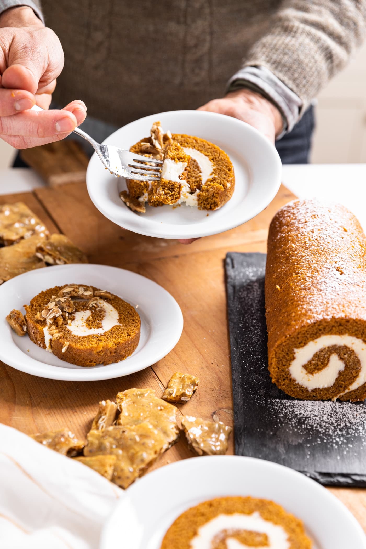 Hand holding white plate filled with slice of pumpkin roll with cream cheese center with other slices sitting around along with rest of the roll