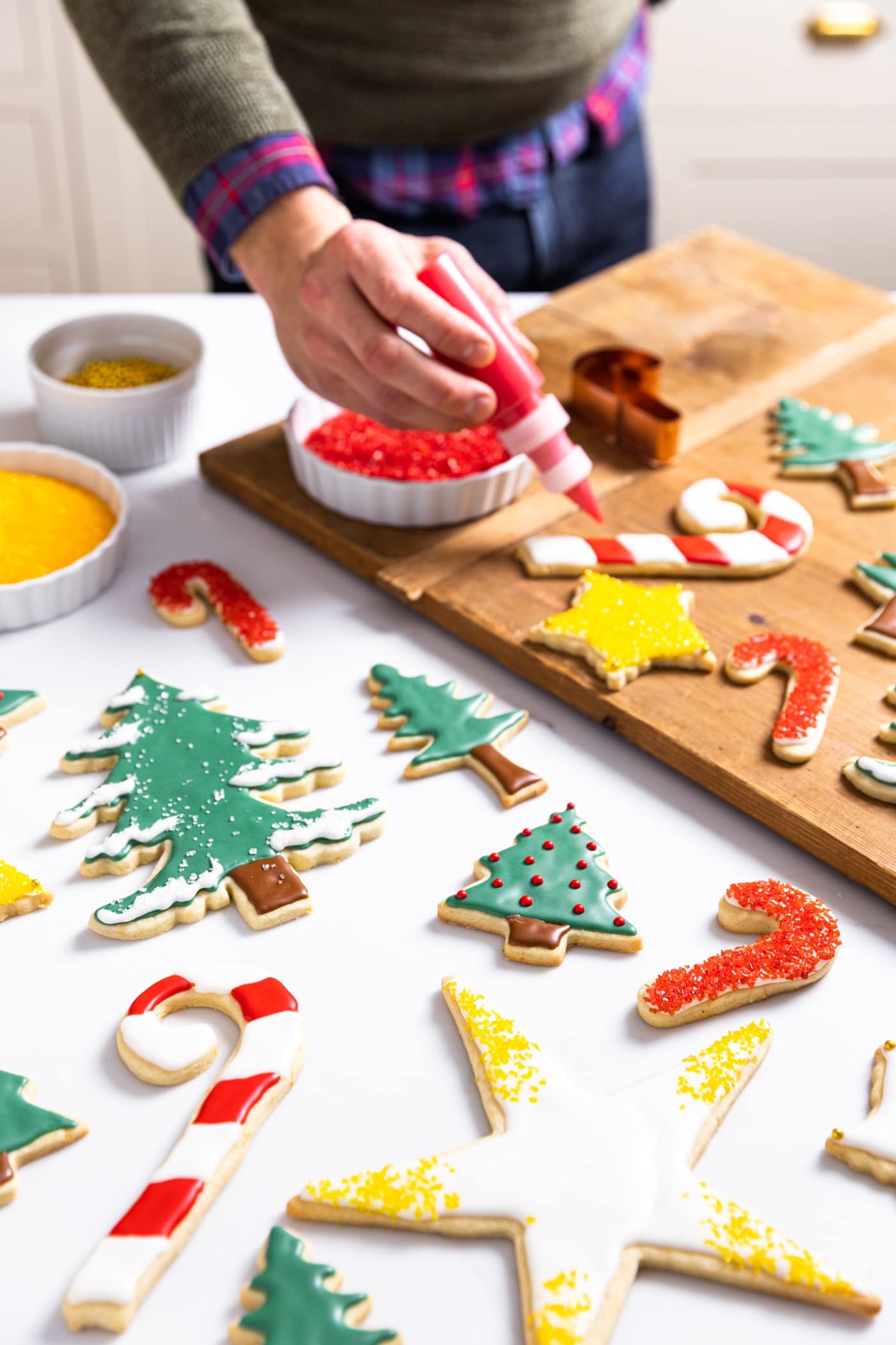 Hand holding squeeze bottle filled with red food coloring decorating Christmas sugar cookies sitting on white countertop