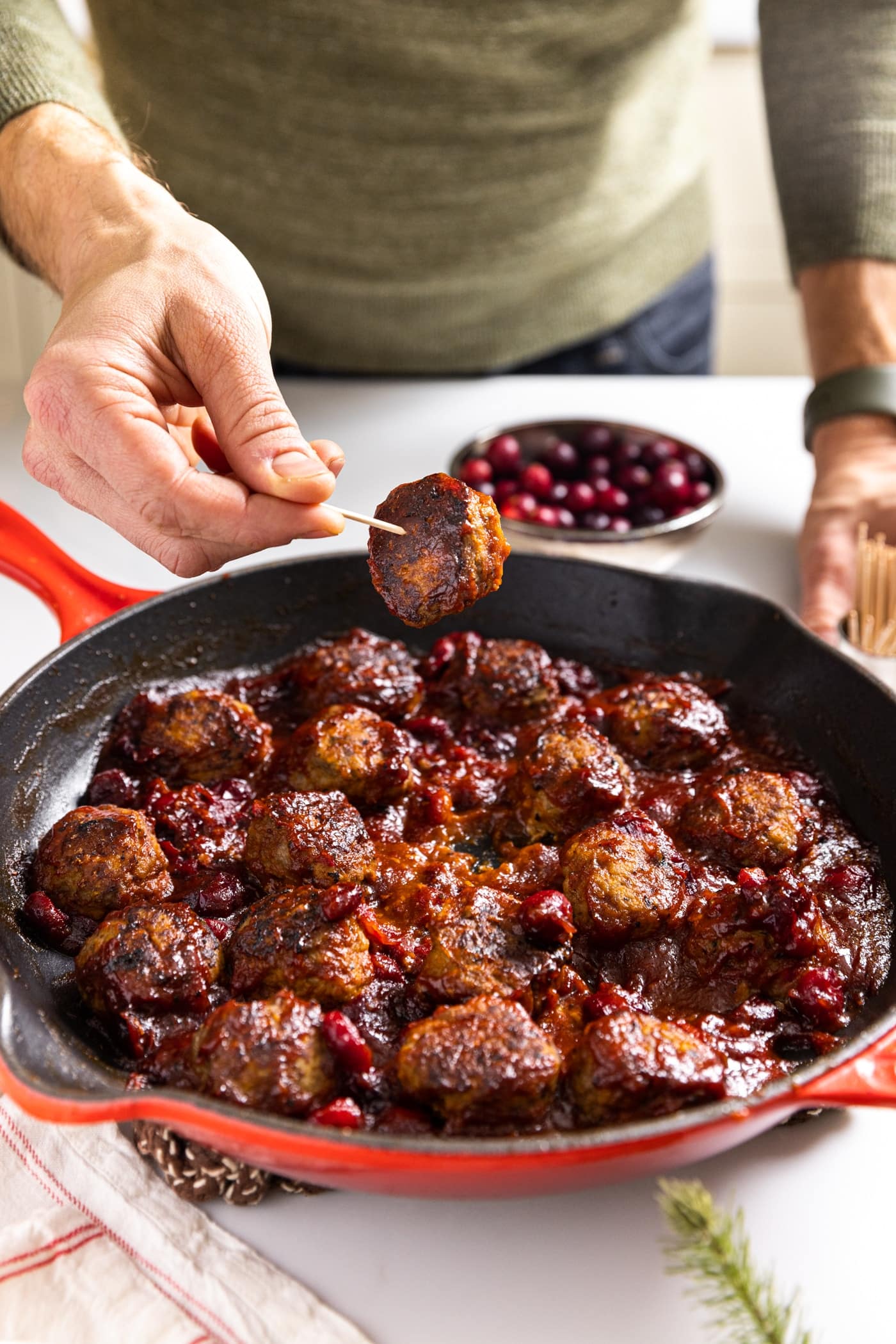 Hand holding wood toothpick speared with small meatball covered in cranberry sauce with skillet filled with more meatballs below all on white countertop