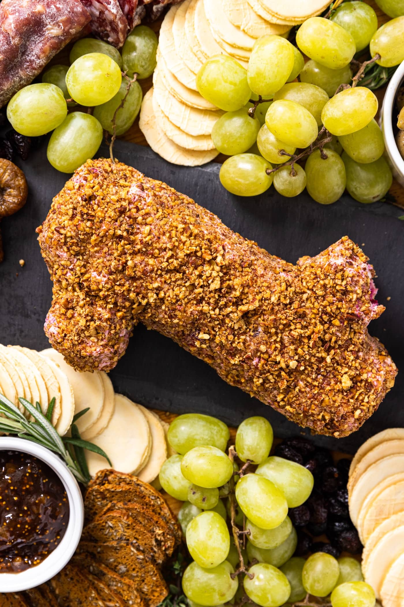 Top down view of piece of slate with cranberry cheese log shaped like a piece of wood covered in crushed nuts with grapes and crackers all around