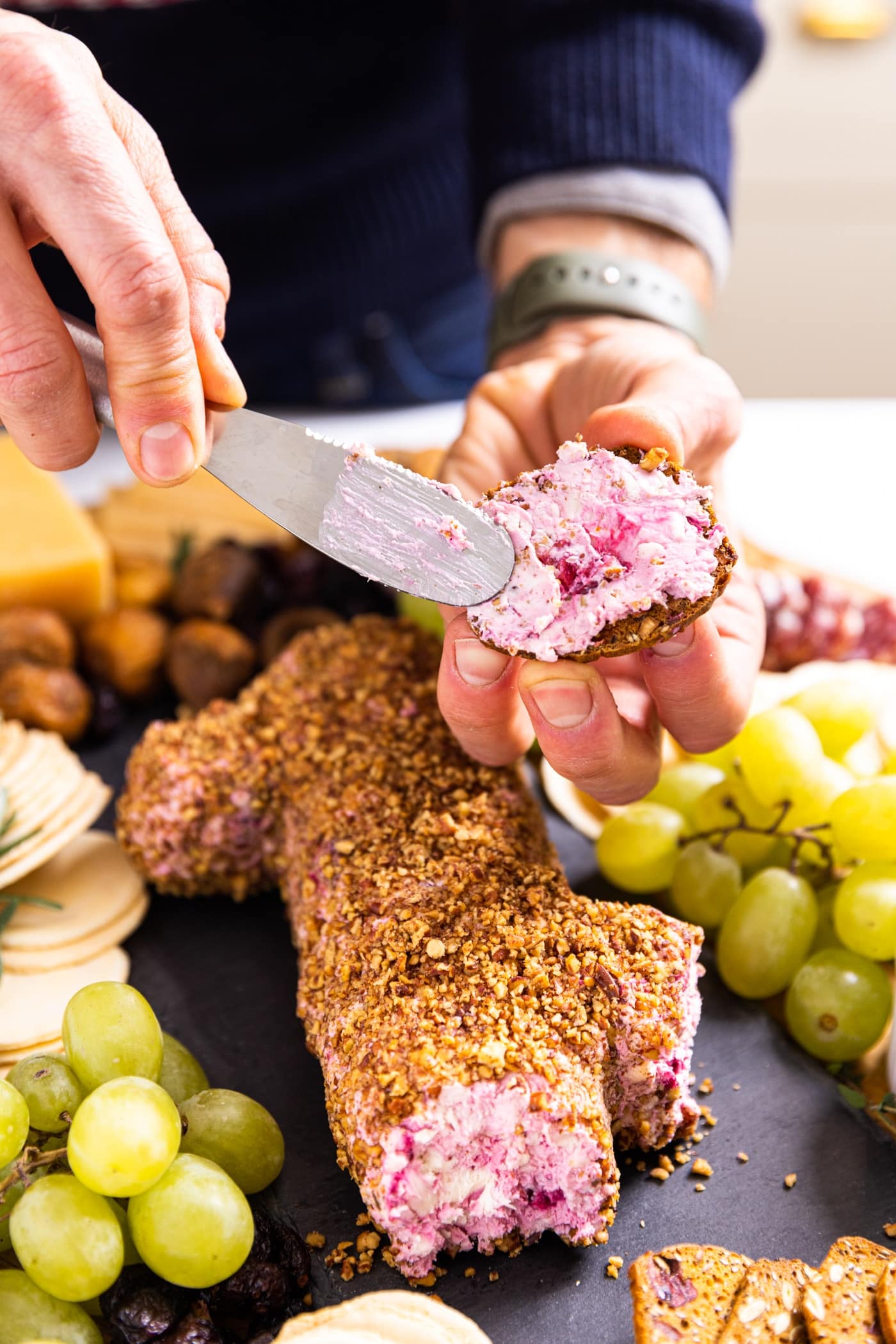Hand holding cheese knife spreading cranberry cheese onto cracker with rest of cranberry cheese log sitting on slate board below