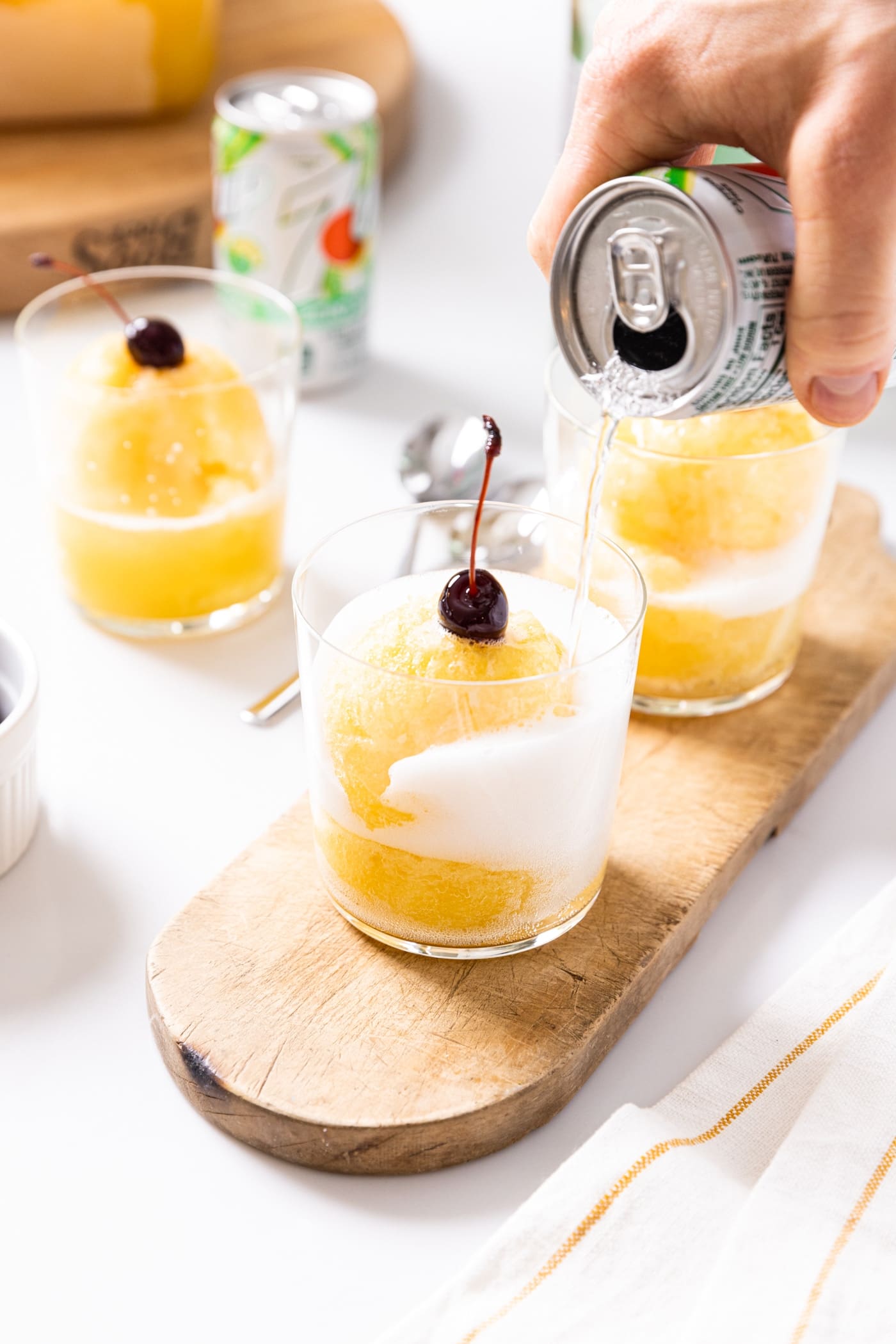 Hand holding soda can pouring soda into clear glass already filled with yellow colored pineapple slush with a cherry on top