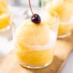 Clear glass sitting on wood board with pineapple slush sitting inside with cherry on top all on white countertop