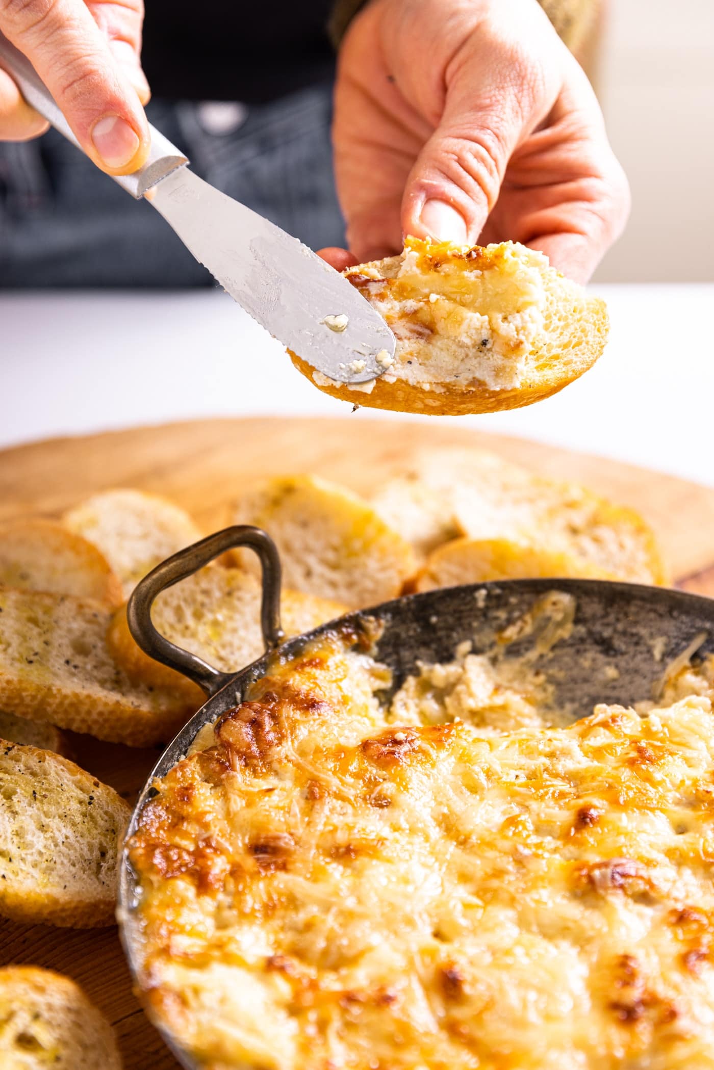 Hand holding piece of crostini using a cheese knife to spread hot cheese onto slice with rest of cheese sitting in bowl below