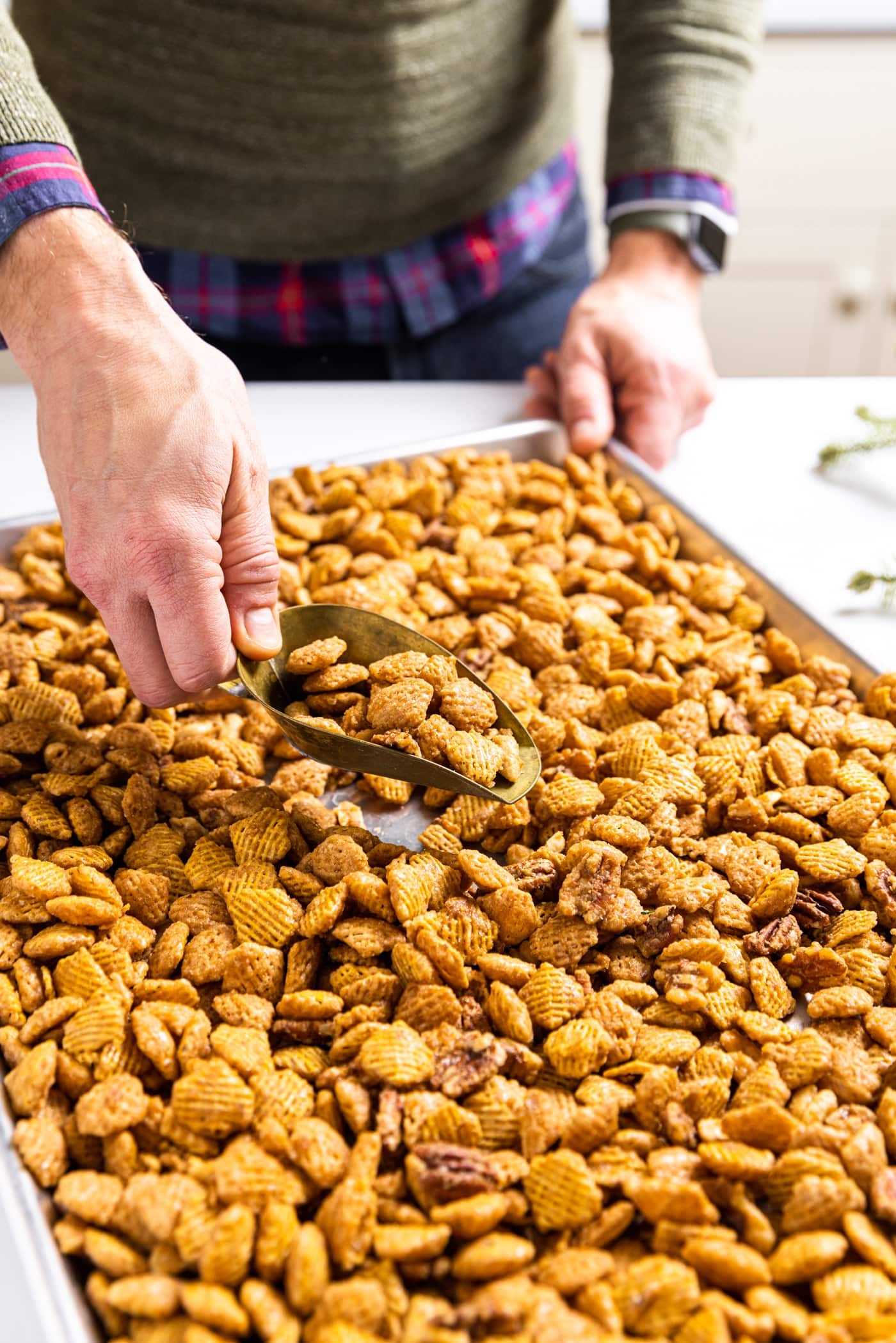 Large metal sheet pan filled with sweet glazed party mix sitting on white countertop with hand holding brass scoop