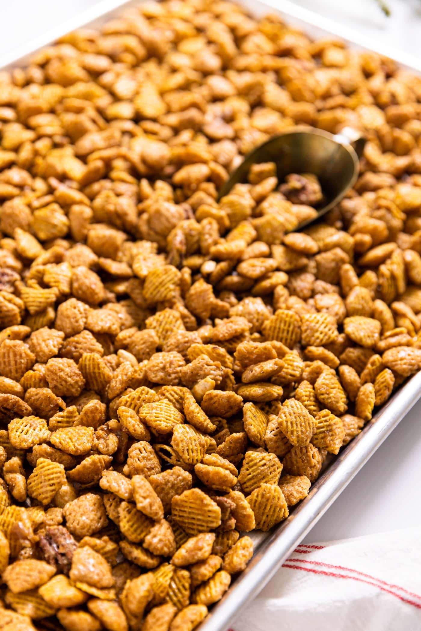 Metal sheet pan filled with yellowish colored glazed party mix with brass scoop scooping out some pieces