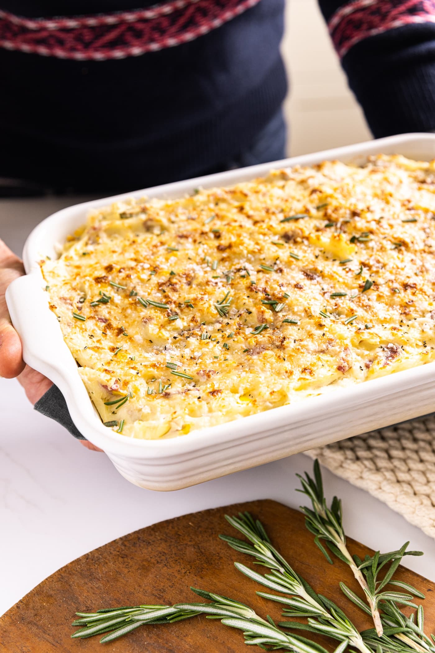 Two hands holding white baking dish filled with smashed potatoes with a golden brown crust on top with wood cutting board with fresh rosemary on top