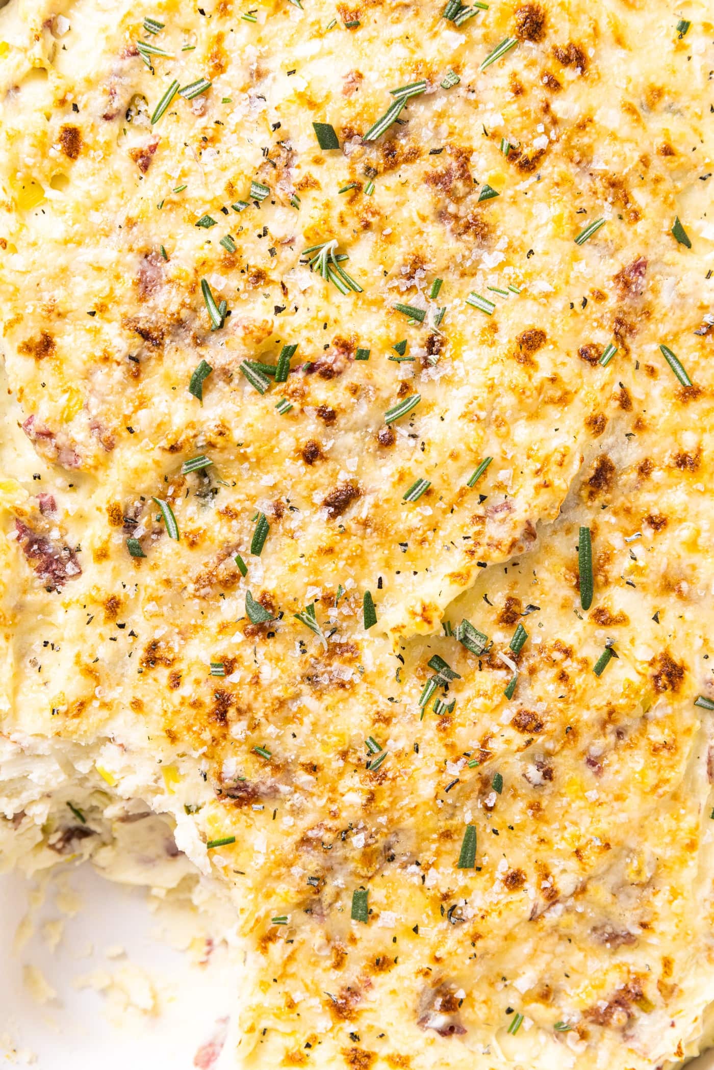 Top down view of golden brown smashed potatoes mixture sitting in white baking dish with fresh minced rosemary and sea salt over the top