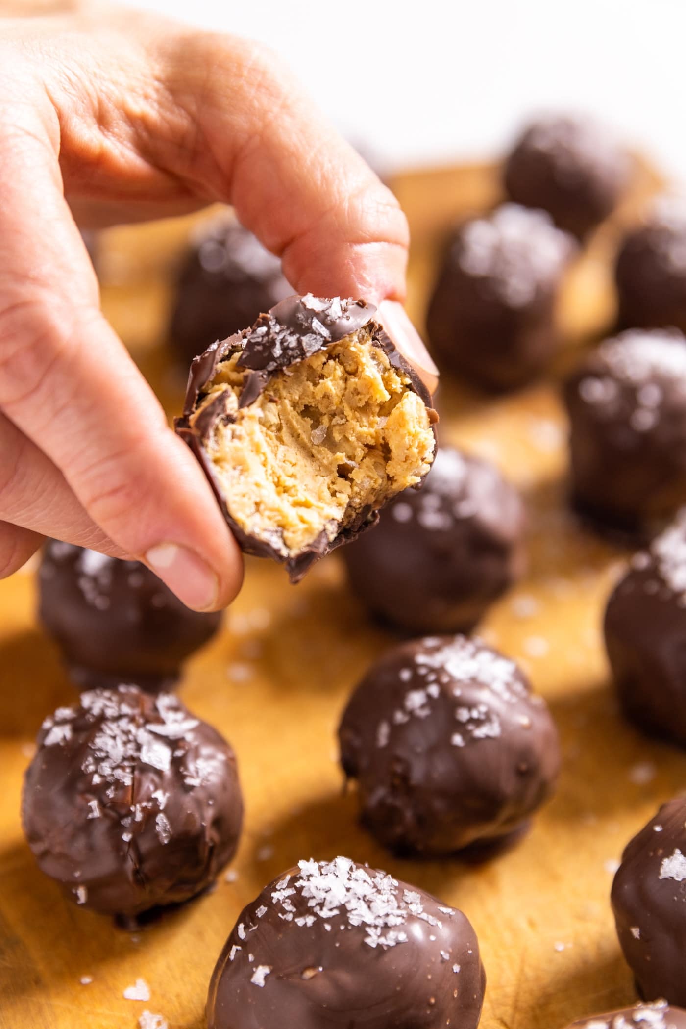 Hand holding peanut butter ball covered in chocolate with bite taken out with other peanut butter balls sitting on wood board