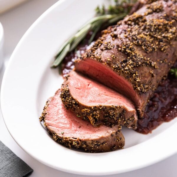 Large white platter with peppercorn crusted beef tenderloin sitting on top with small container filled with pepper